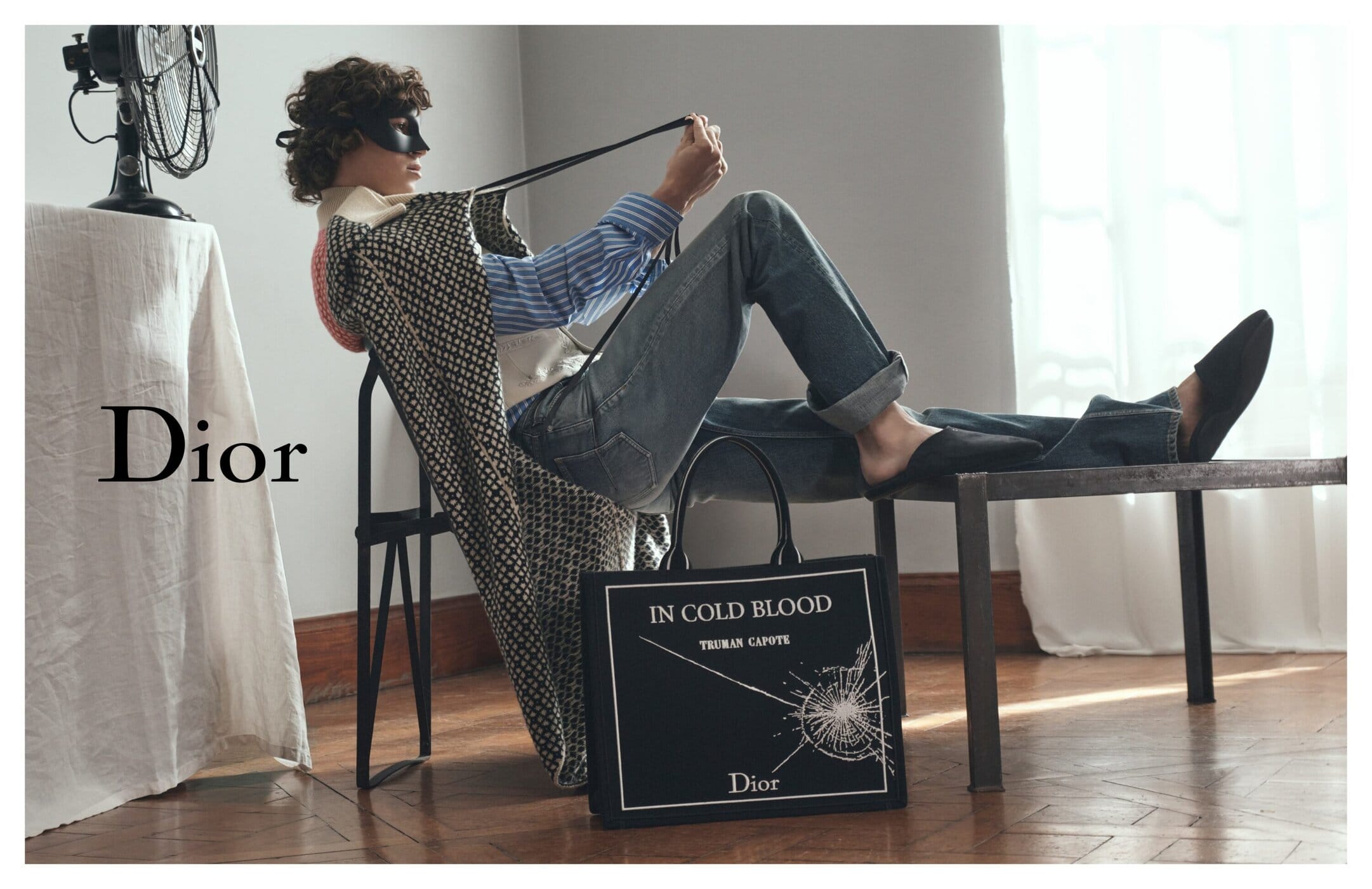 Model wearing a black mask and patterned coat reclining on a stool with a black graphic tote bag on wooden flooring.