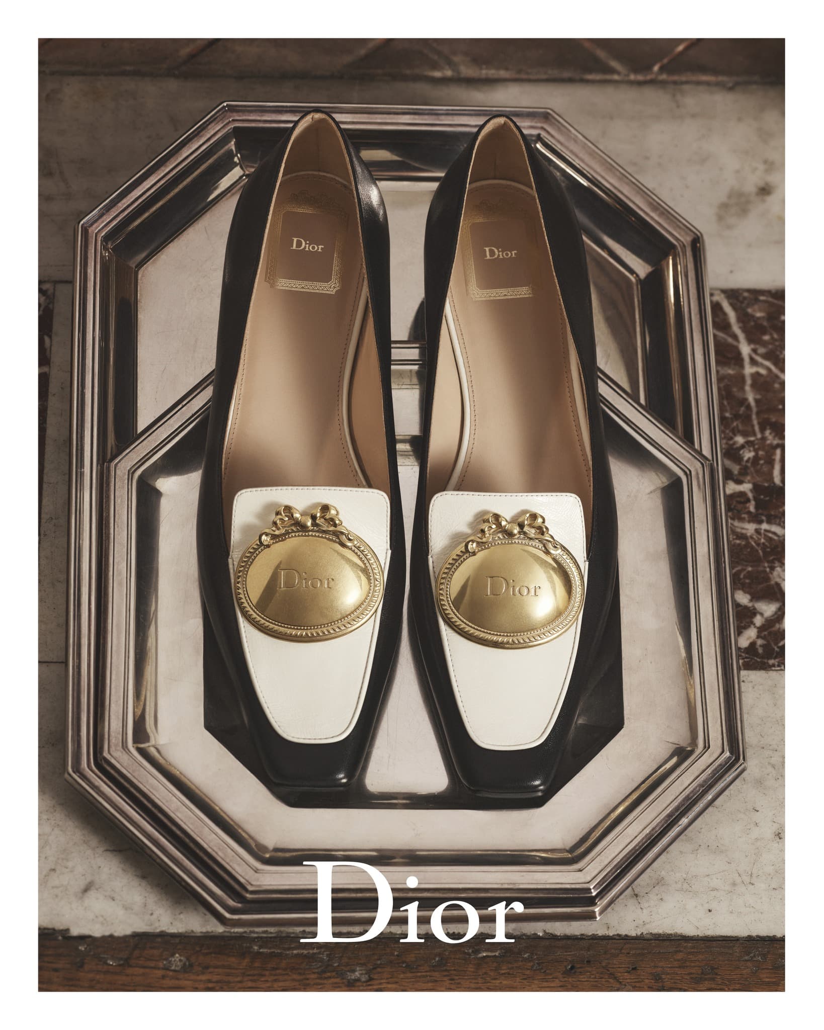 Elevated shot of black and white Dior loafers with gold embellishments, presented on an octagonal silver tray against a marble backdrop.