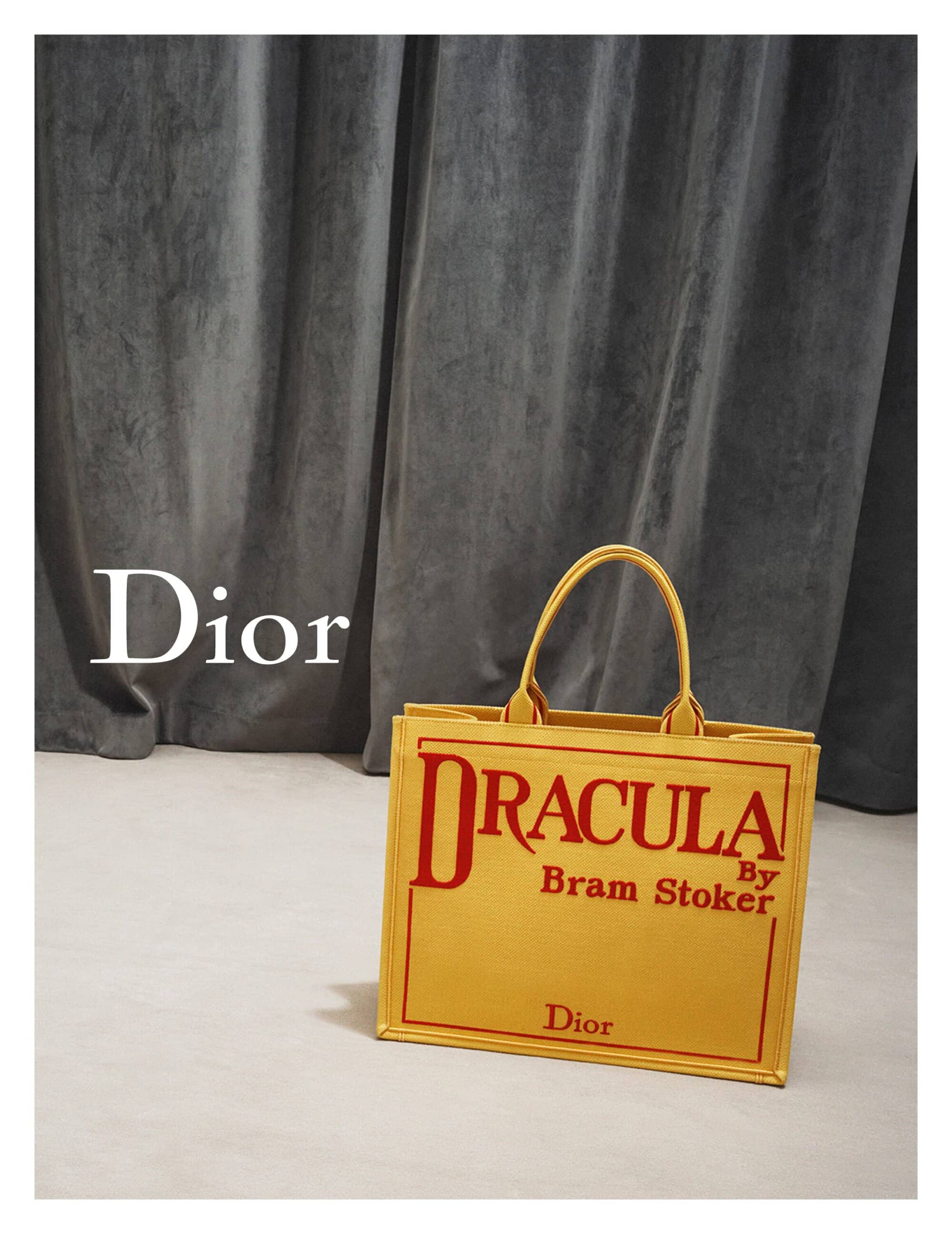 Yellow Dior tote bag with 'Dracula by Bram Stoker' printed in red, against a draped gray velvet backdrop. Fashion product shot.