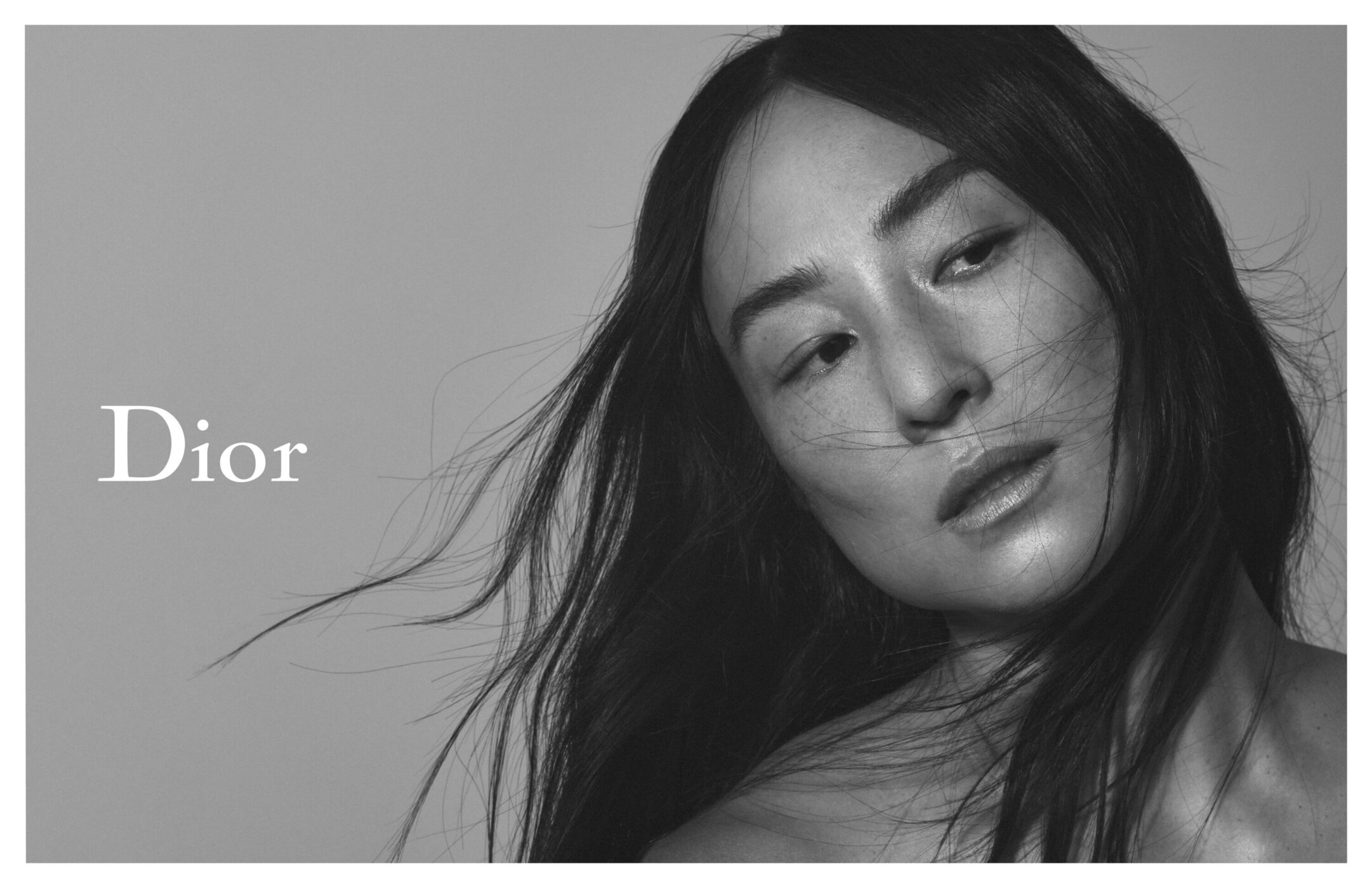 Close-up studio shot of a woman with flowing dark hair and neutral makeup looking down, featuring Dior branding on a grey background.