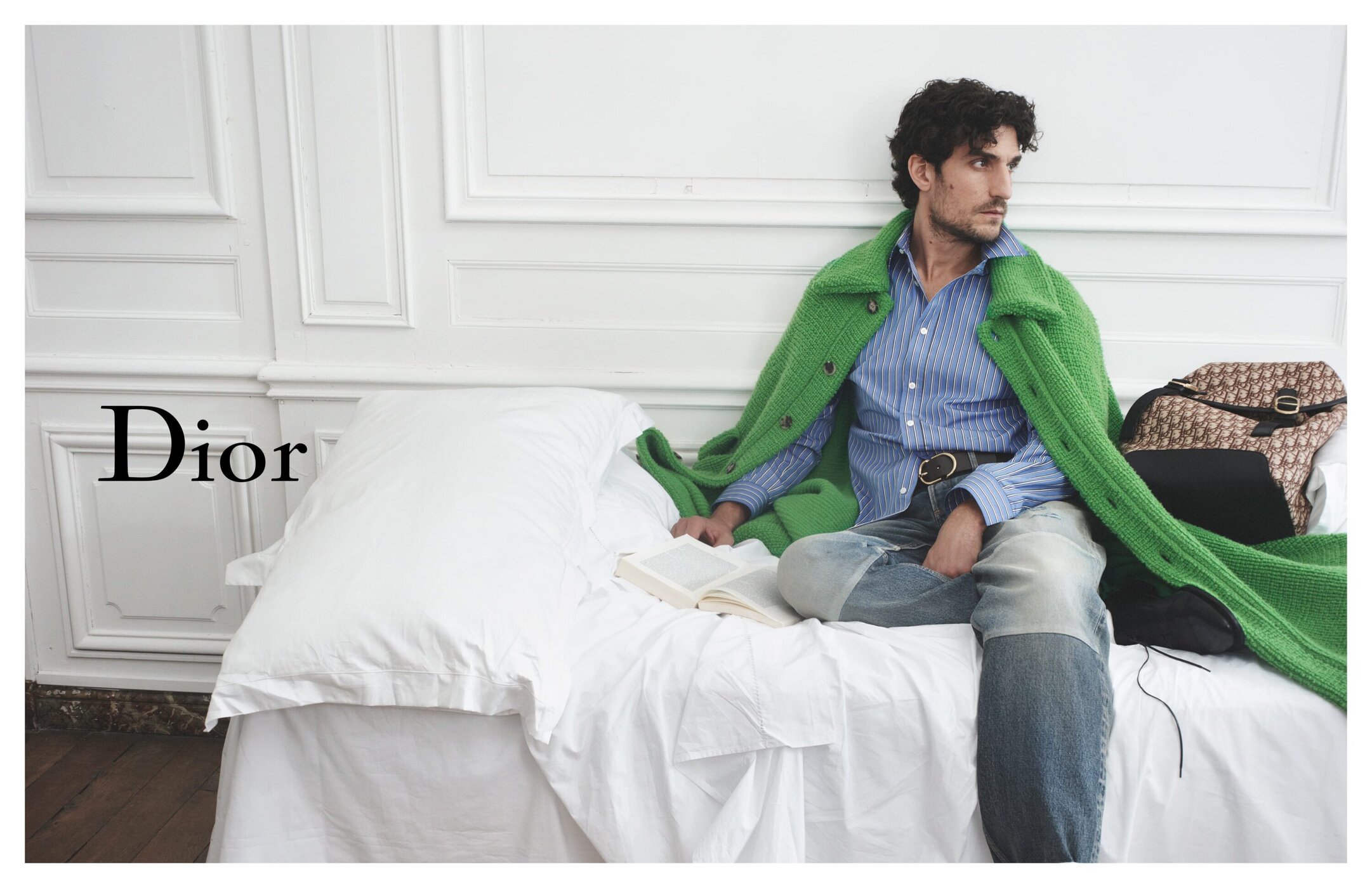 Adult male model sits on a white bed in an indoor setting, draped in a bright green jacket and holding a book with a Dior bag nearby.