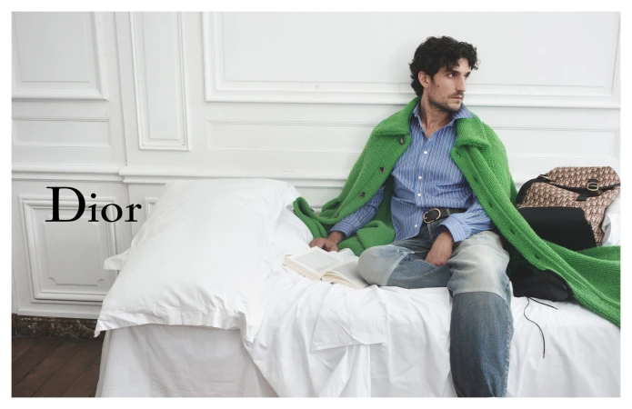 Adult male model sits on a white bed in an indoor setting, draped in a bright green jacket and holding a book with a Dior bag nearby.