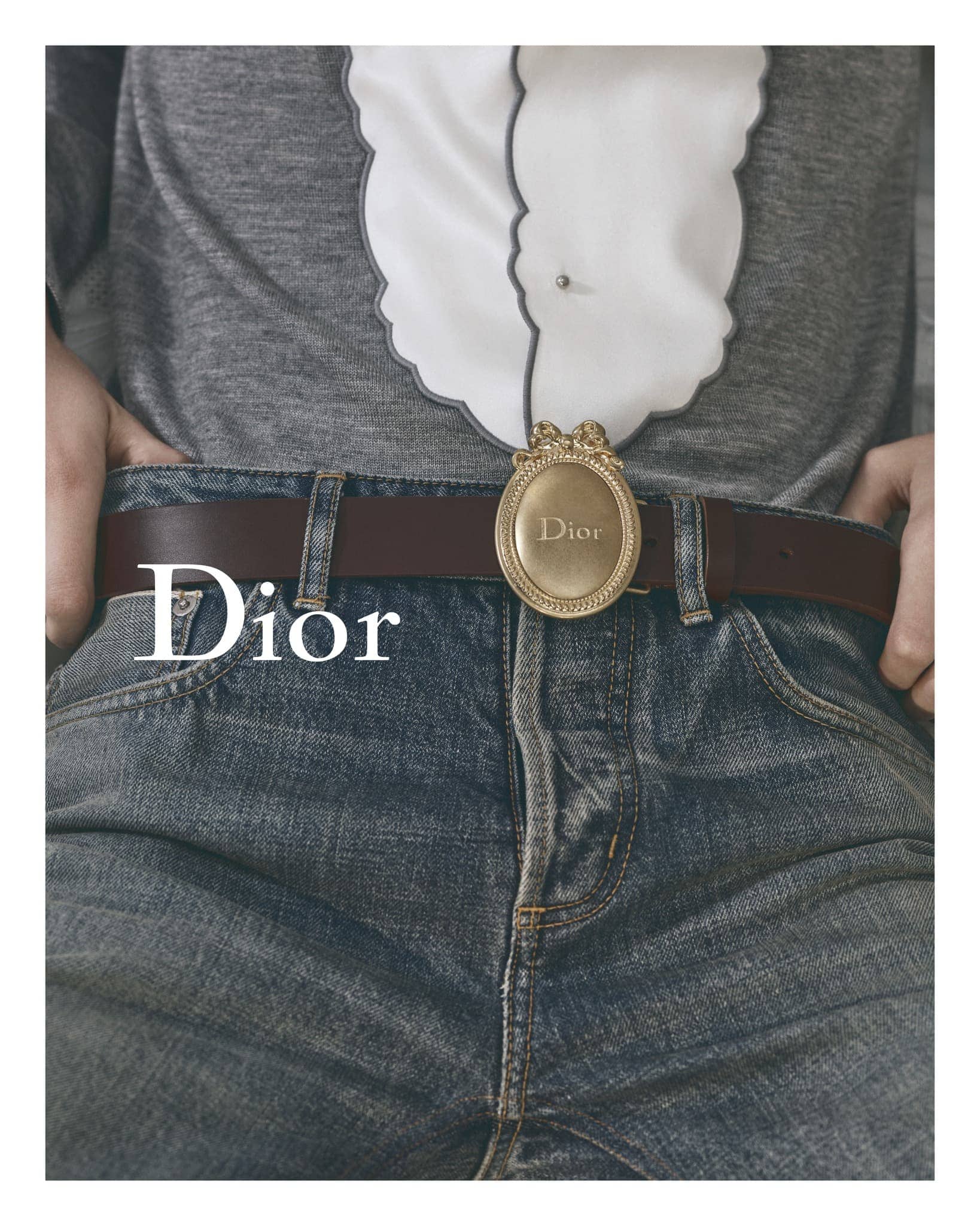 Waist-up shot showcases blue jeans, brown leather belt with a gold Dior buckle, worn with a gray sweater and white frilly shirt.