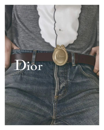 Waist-up shot showcases blue jeans, brown leather belt with a gold Dior buckle, worn with a gray sweater and white frilly shirt.