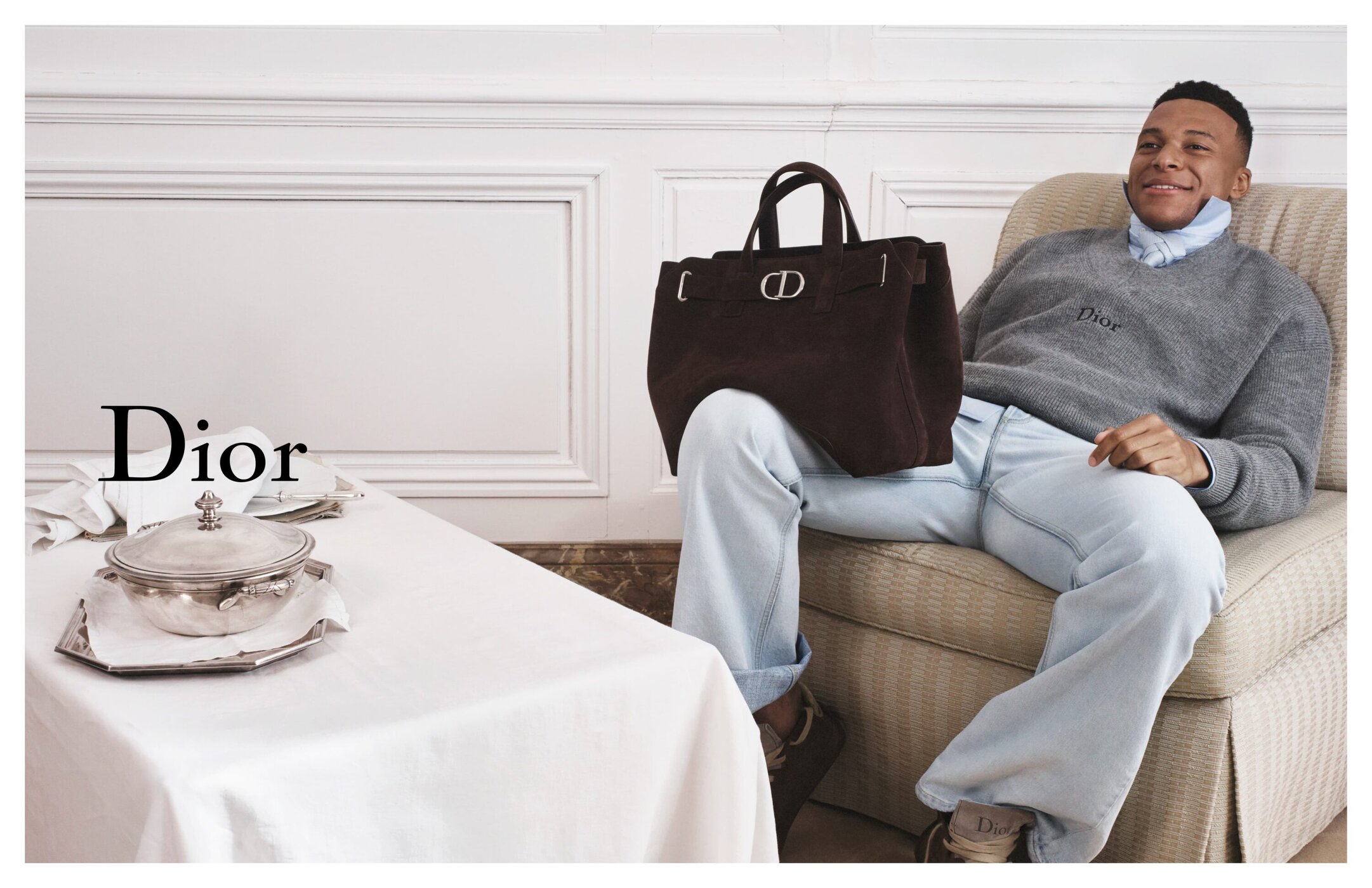 A man sits on a beige armchair with a brown designer purse on his lap, in a white room. Table with Dior branded pot in foreground.