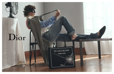 Model wearing a black mask and patterned coat reclining on a stool with a black graphic tote bag on wooden flooring.
