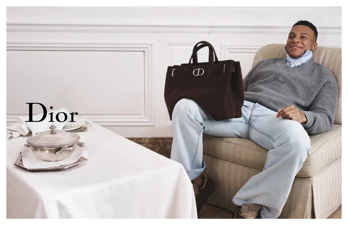 Kylian Mbappe seated in an armchair holding a dark brown suede handbag for Dior Spring 2026 campaign.