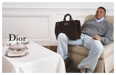 Kylian Mbappe seated in an armchair holding a dark brown suede handbag for Dior Spring 2026 campaign.