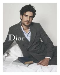 Louis Garrel wearing a grey double breasted wool suit and white shirt reclining on white bedding with two books for Dior