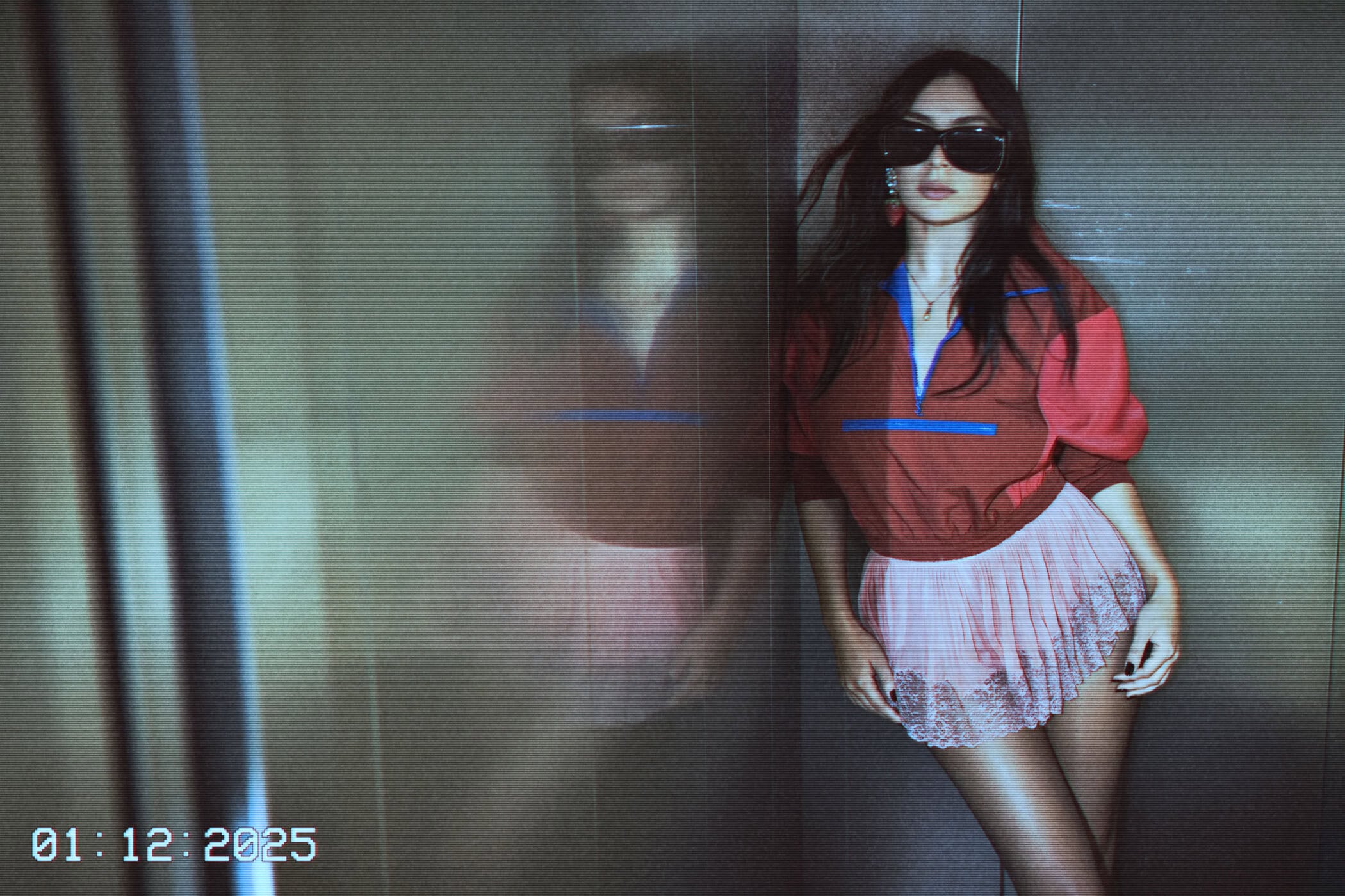 A stylish woman with sunglasses poses in an elevator. She wears a colorful cropped top and pink skirt, leaning against the silver wall.