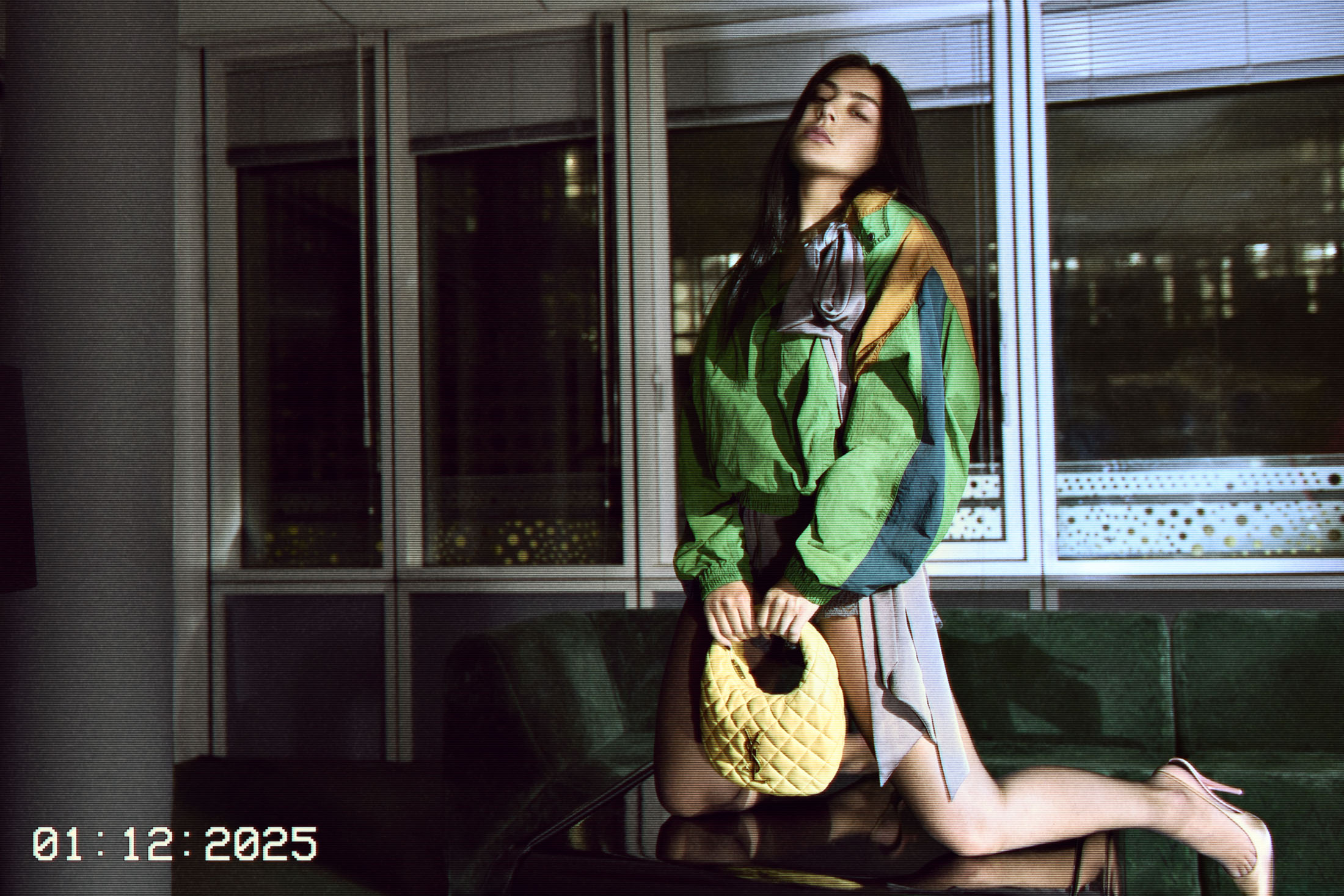 Woman in multi-colored jacket kneels on couch holding yellow handbag with windows in the background. Fashion editorial shot, indoor setting.