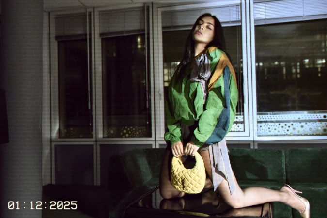 Woman in multi-colored jacket kneels on couch holding yellow handbag with windows in the background. Fashion editorial shot, indoor setting.
