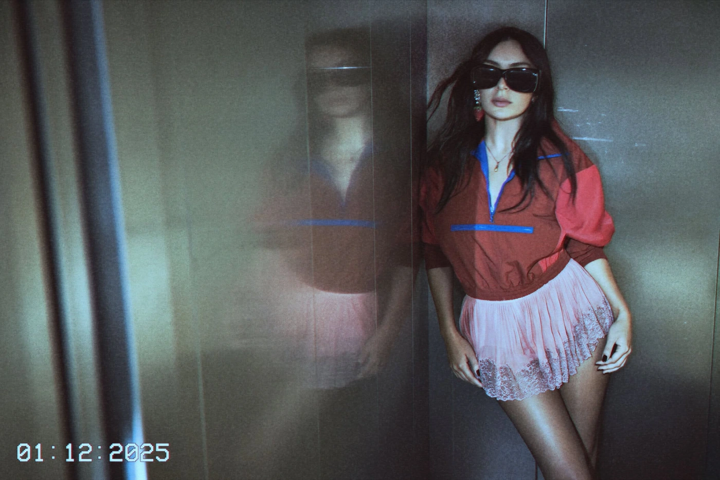 A woman wearing a red pullover and pink skirt leaning against a metallic wall with reflections, shot by Glen Luchford for Saint Laurent.