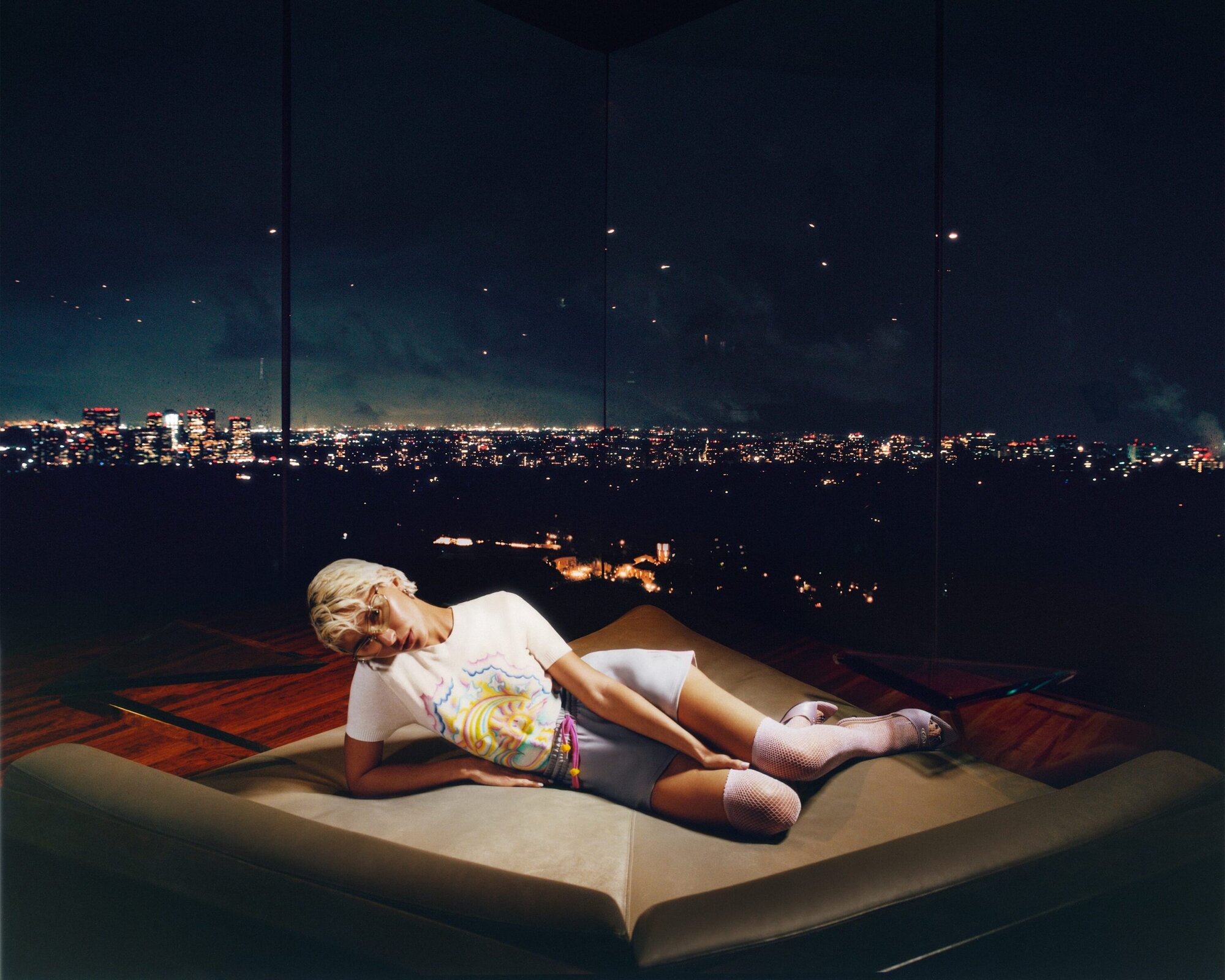 Nighttime view of a woman with blonde hair reclining on a lounge chair, illuminated by city lights through floor-to-ceiling windows.