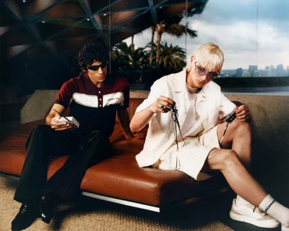 Two men sit on a brown couch in an architectural interior, wearing sunglasses and casual clothing while holding audio devices.