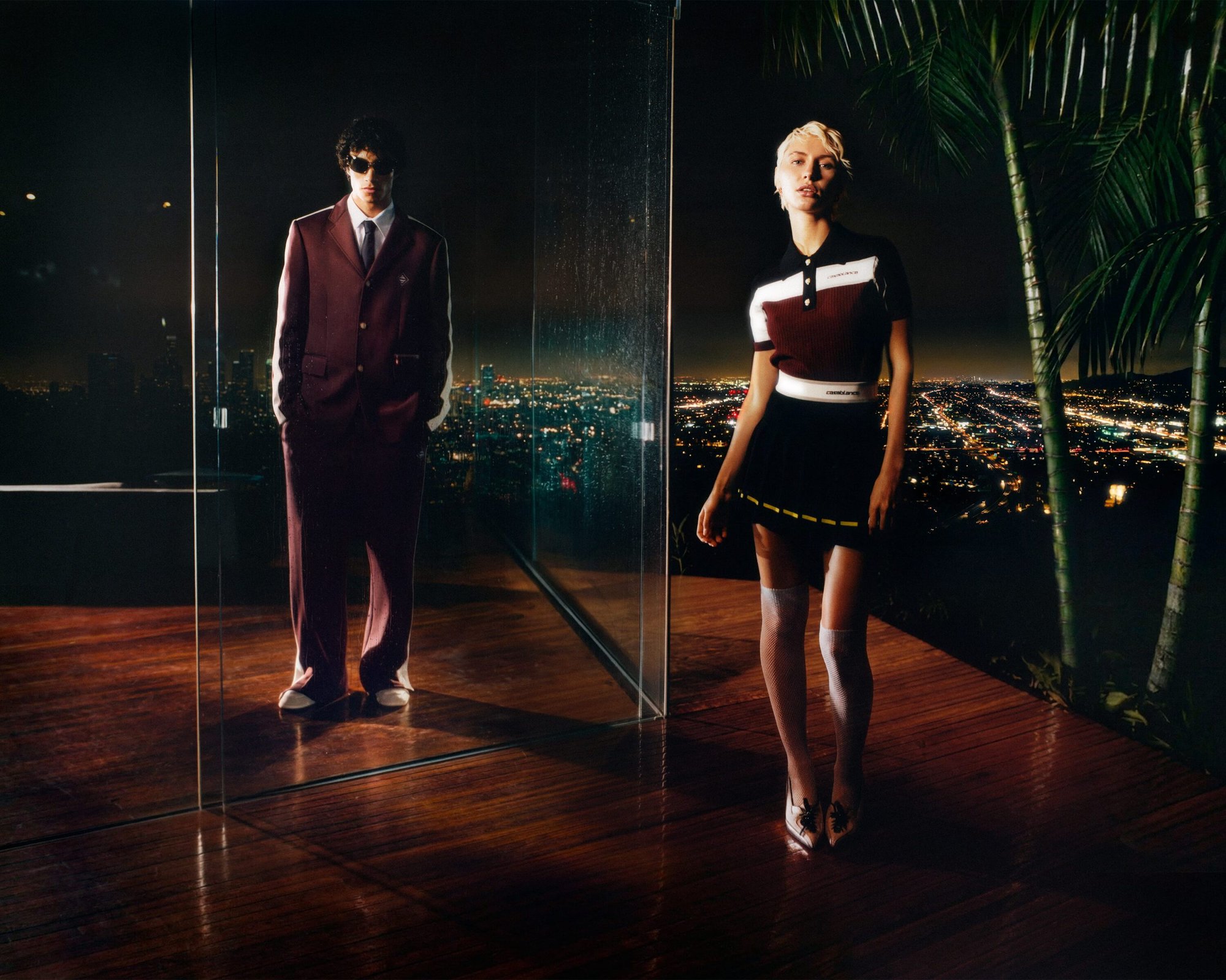 Two fashion models posing in front of a city skyline at night; an adult male is wearing a suit, and an adult female is wearing a skirt and top.