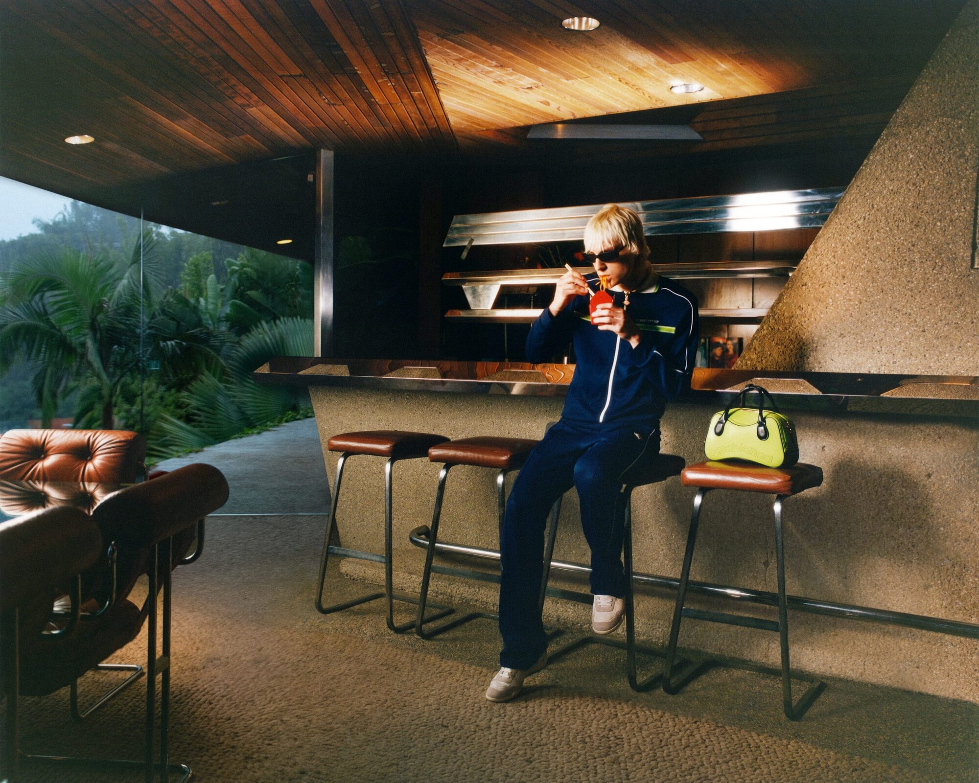 Woman in blue tracksuit sits on a bar stool indoors, holding a red drink with straw next to a green purse in front of a bar.
