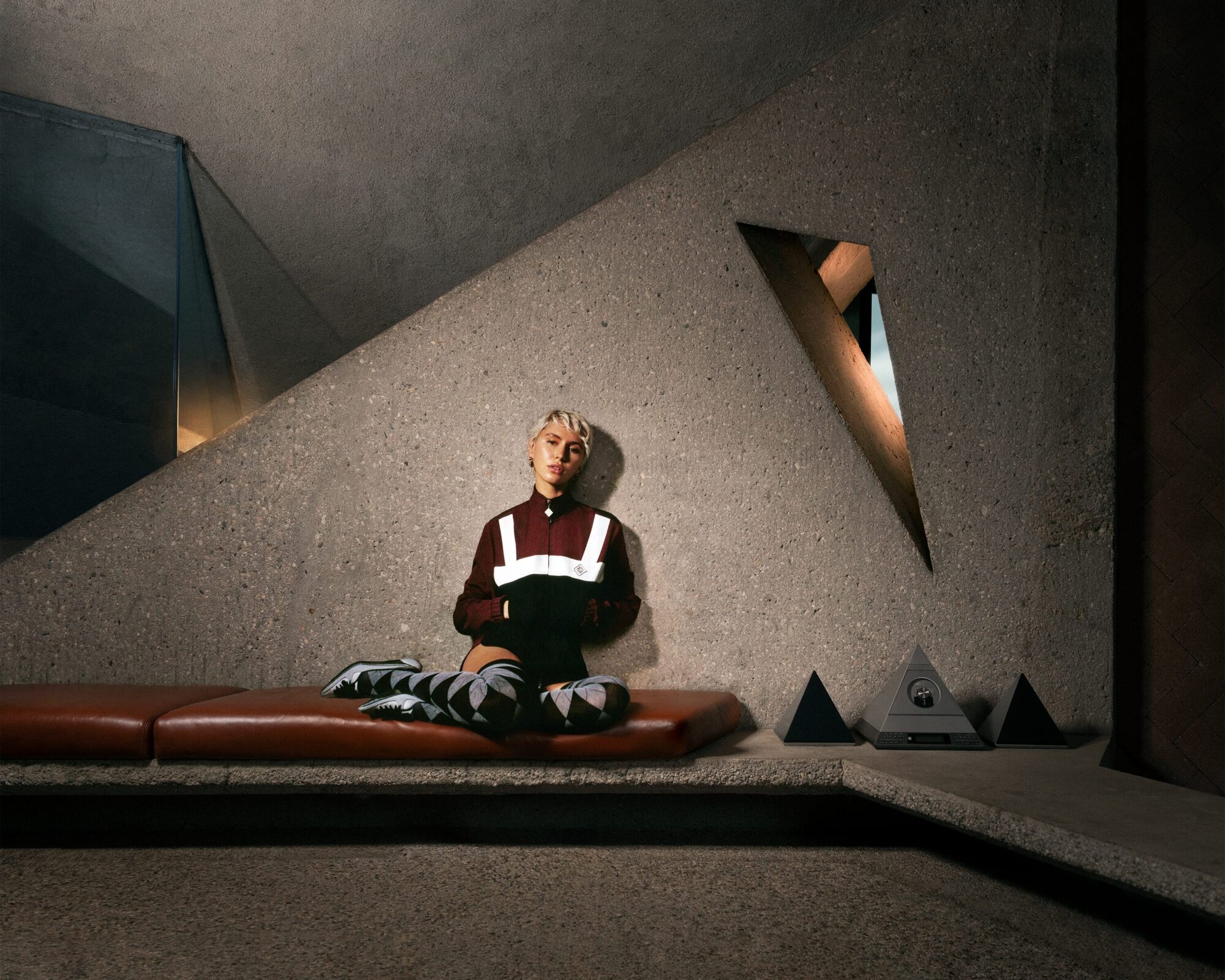 Woman with short blonde hair sits cross-legged on a leather bench in an angular concrete interior with pyramidal decor and accent lighting.
