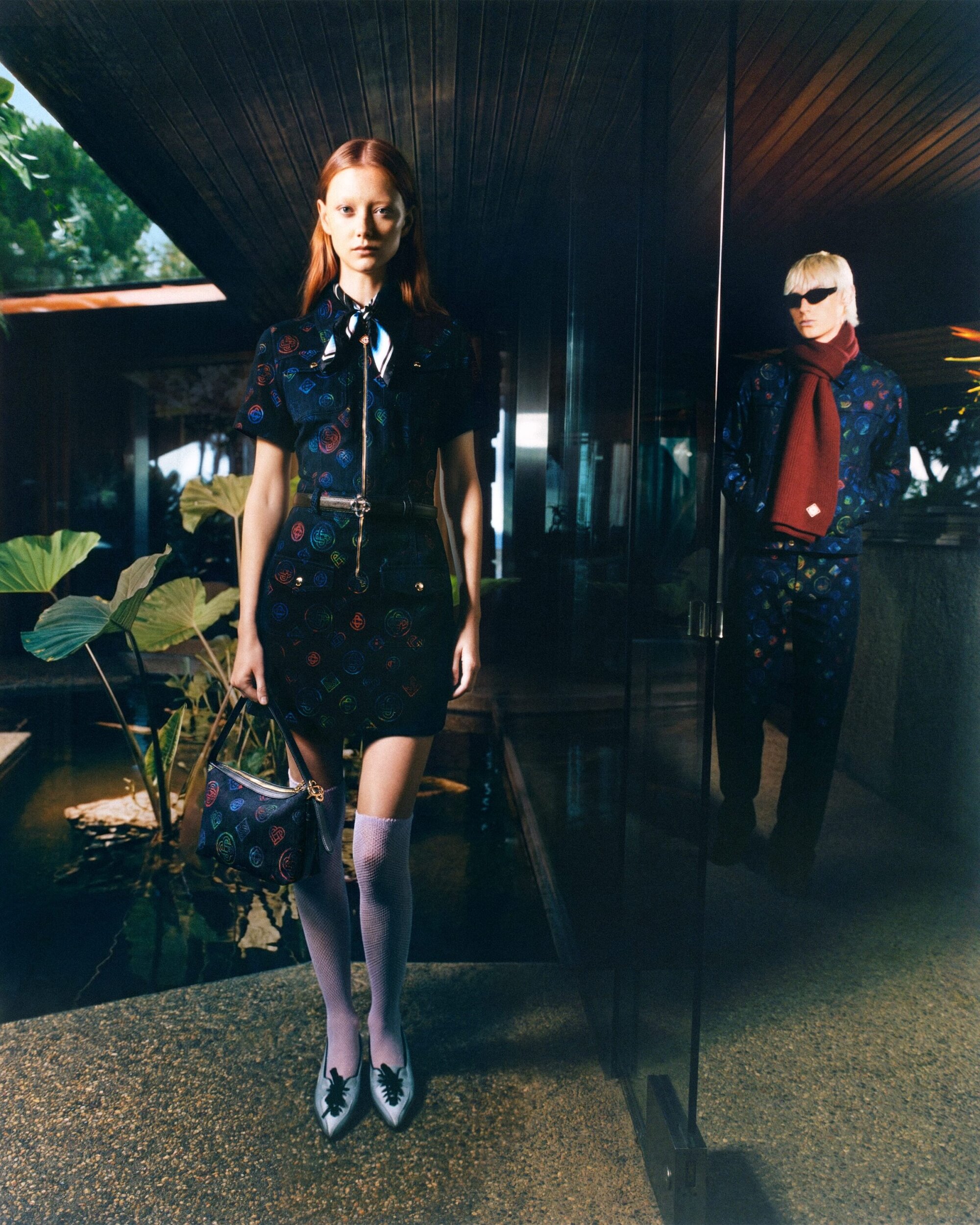 Two fashion models pose outdoors in patterned dresses and hosiery near a reflective pool, conveying an editorial style and luxury fashion.