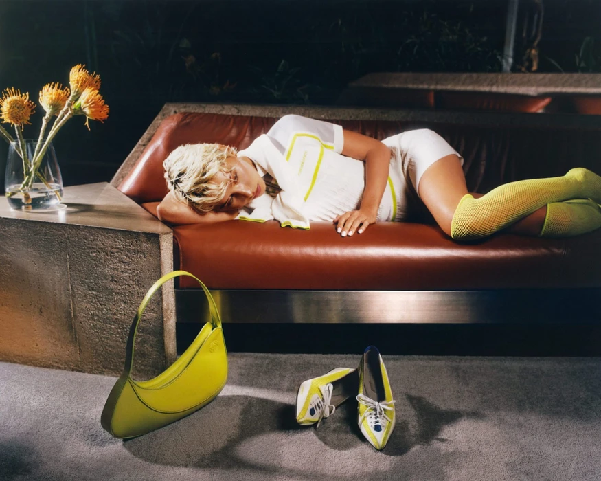 Model with blonde hair reclining on a dark leather sofa beside a vase of orange flowers, with a yellow handbag and shoes on the floor.