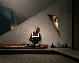 Model seated on a leather bench against a brutalist concrete wall wearing a maroon jacket and patterned over-the-knee socks.