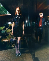 Model in patterned dark dress and knee-high socks holding handbag with another person standing in background