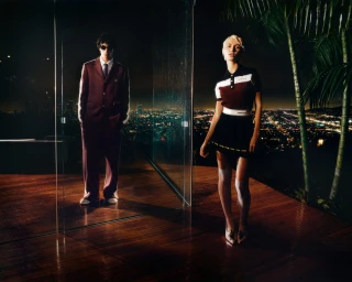 Man in maroon suit standing behind glass and woman in short-sleeved top and skirt on outdoor deck overlooking night city lights