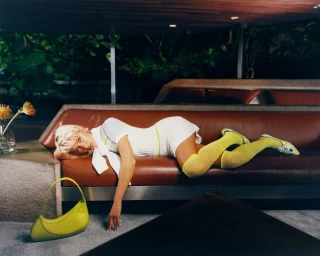 Model in white dress and bright green socks lying on brown leather sofa in Casablanca Resort 2026 campaign.
