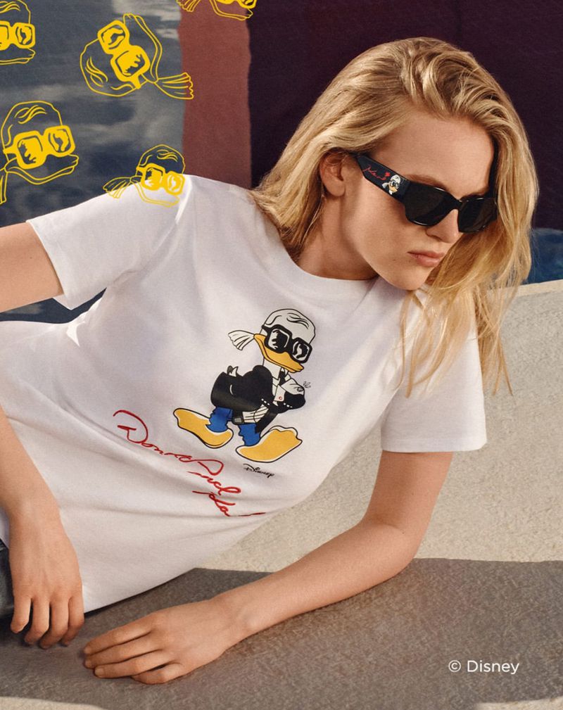 Adult female wearing sunglasses and a white t-shirt with a Donald Duck cartoon lying in an outdoor setting on a sunny day.