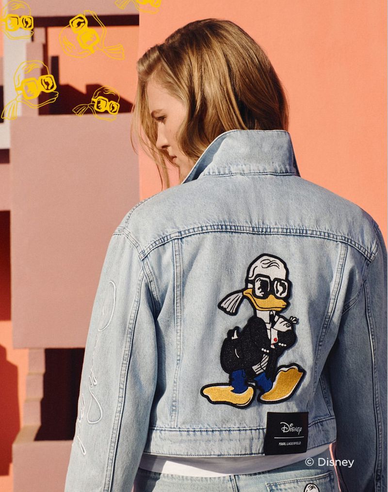 A fair-skinned woman models a light denim jacket with a cartoon graphic on the back, set against a pink and white background.