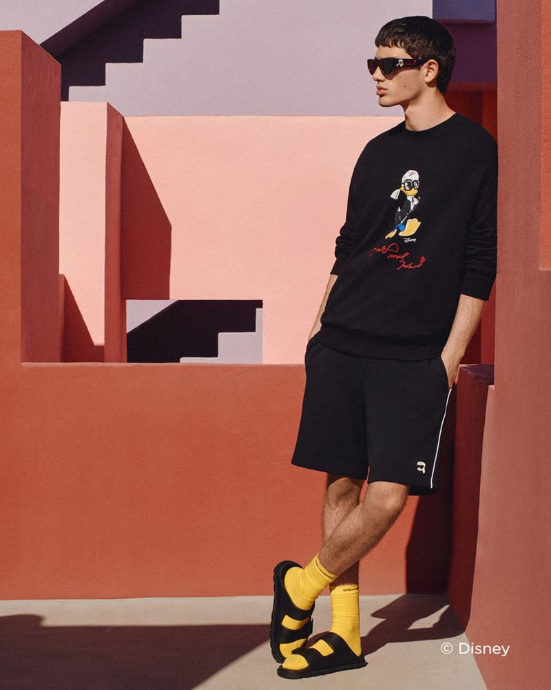 Man in black Karl Lagerfeld sweatshirt and shorts leaning against a peach wall, wearing sandals and yellow socks, sunglasses, studio shot.