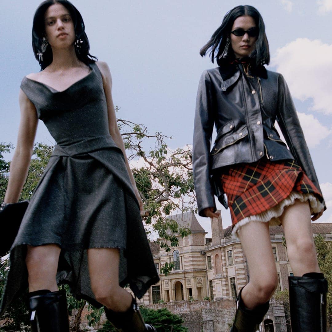 Two women pose outdoors in fashion outfits; one in a grey dress and boots, the other in a black jacket, plaid skirt, and black boots.