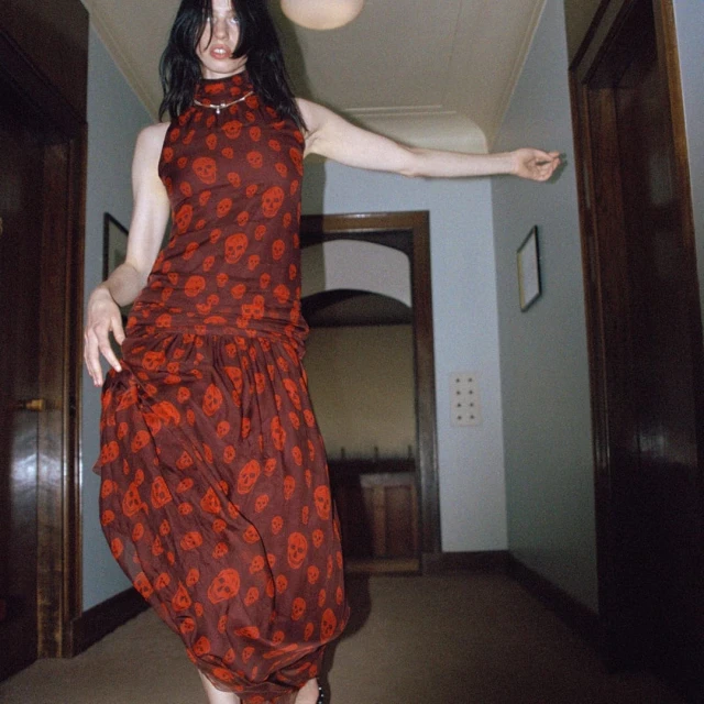 A woman in a long, dark red dress with a skull print stands in a hallway with a slight smile, posing with one arm extended.