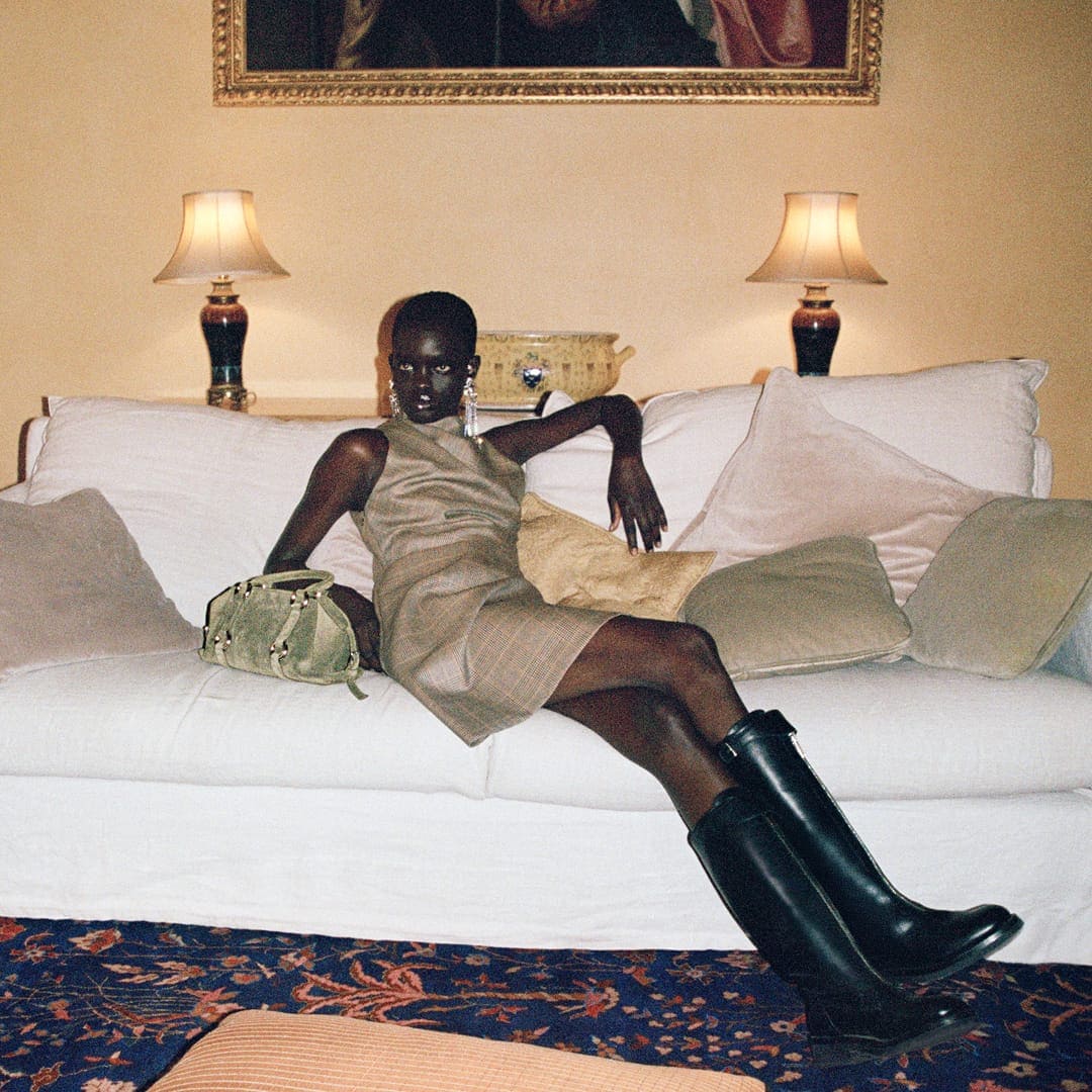 Elegant woman in dress and boots poses on a white sofa with pillows, with a handbag on the sofa and painting on the wall.