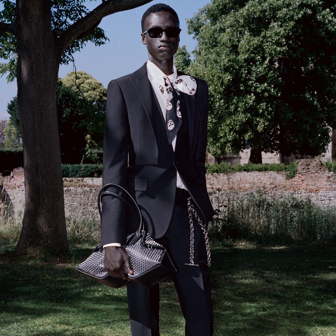 A man wearing a black suit and sunglasses holds a studded handbag in a park setting.