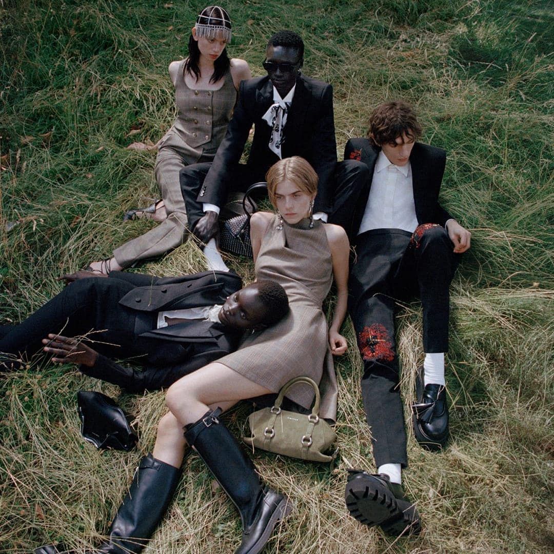 Five fashion models sit in a grassy field wearing stylish outfits. Diverse group poses in black suits, a dress, and a vest-and-pants combo.