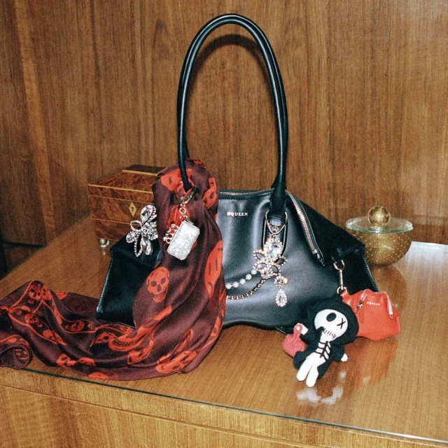 Alexander McQueen black handbag, burgundy skull-print scarf, and charm accessories sit on a brown wood surface against a wood wall.
