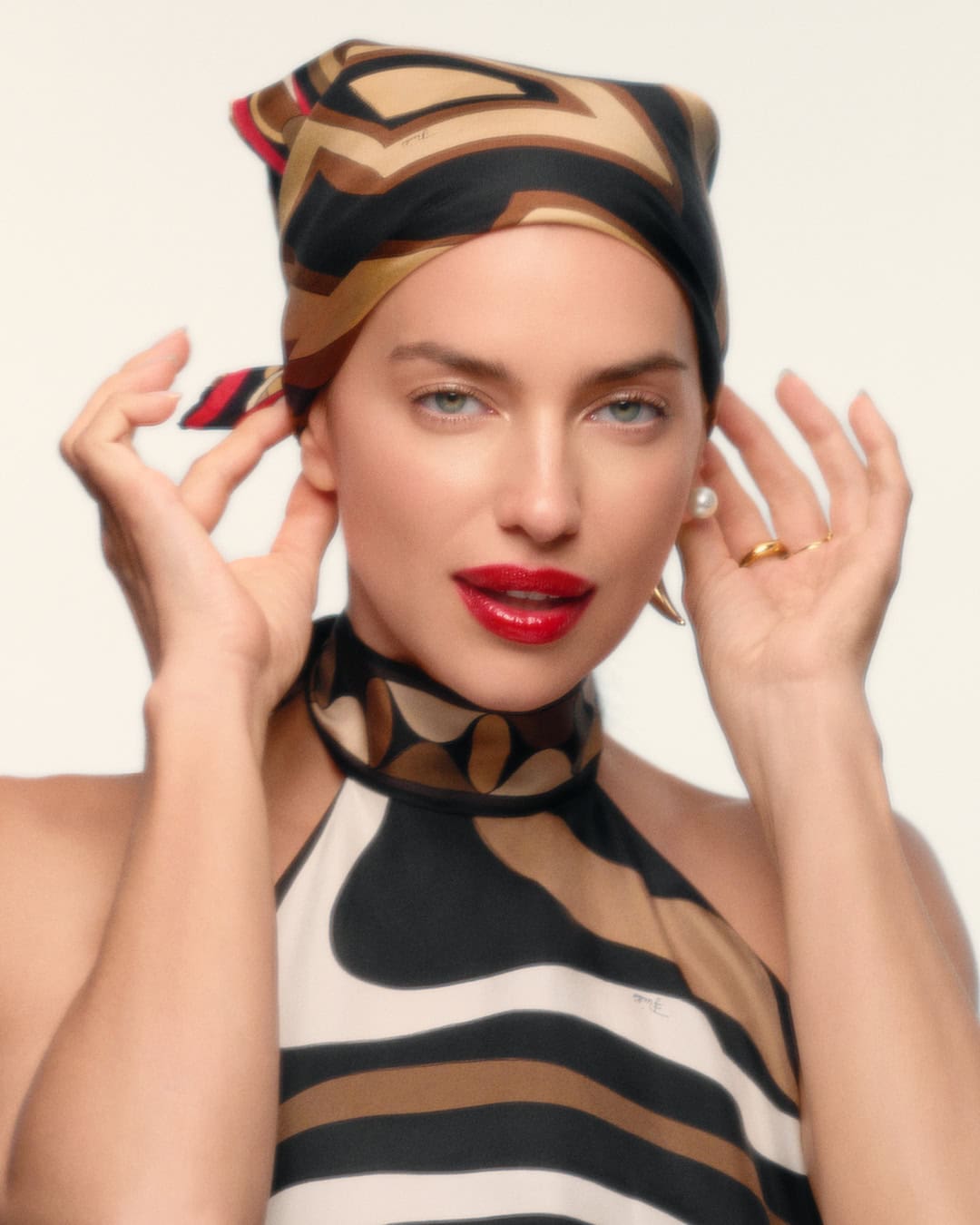Close-up of a woman with red lipstick wearing a patterned headscarf, pearl earrings, and a halter top against a white background.