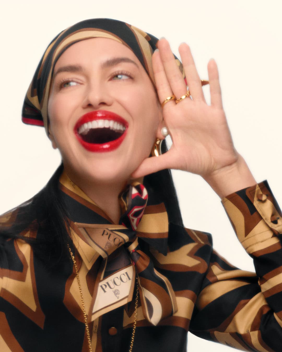 Headshot of adult woman with an open mouth and hand near face wearing a headscarf and blouse with a geometric pattern and pearl earrings.