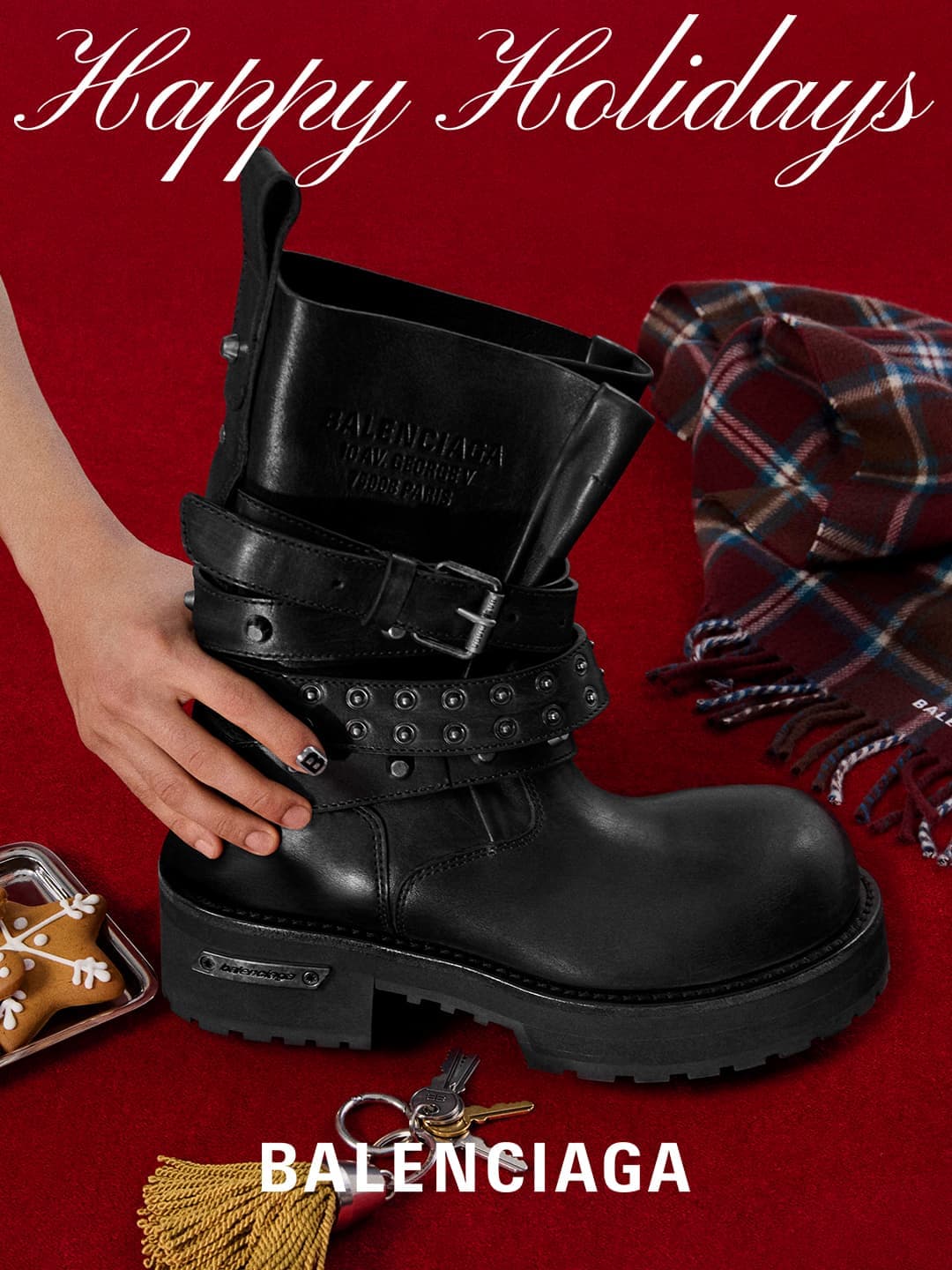 Black Balenciaga boot with straps, silver hardware, and keys, on a red background with cookies and a plaid scarf for holiday fashion.