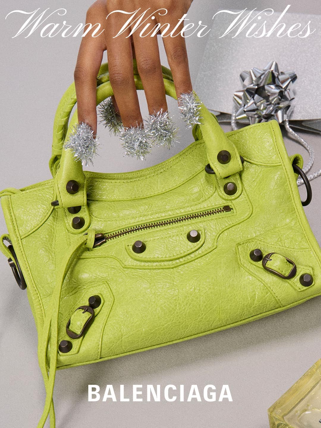 Close-up of a light-green Balenciaga handbag with a fair-skinned hand showcasing silver tinsel nail decorations, with a gift in the background.