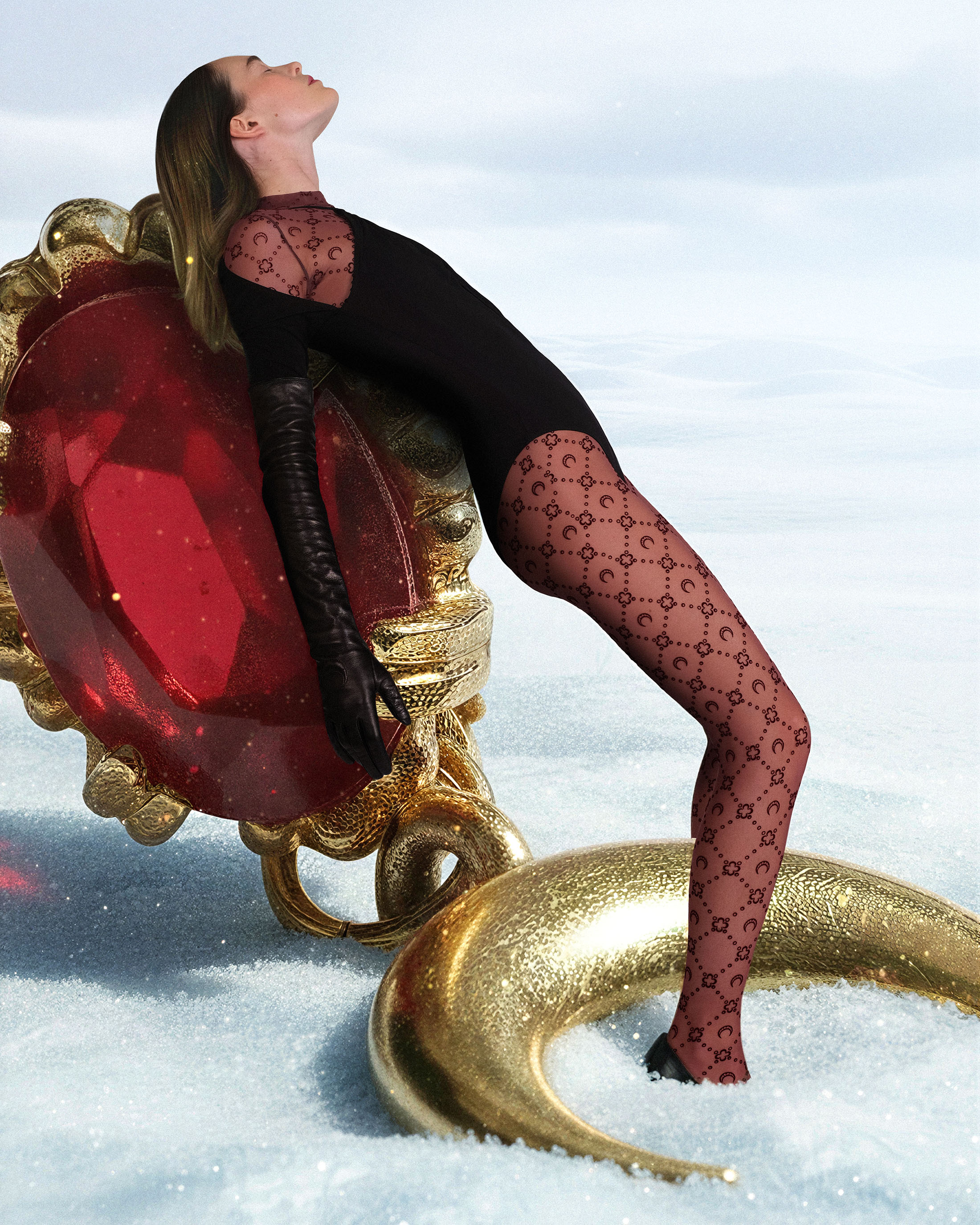 Woman in black bodysuit and patterned tights leaning back on an ornate golden and ruby prop against a sky and snow background.
