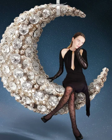 Woman in black dress and black gloves poses while sitting on a bejeweled moon prop with a star-patterned night sky in the background.