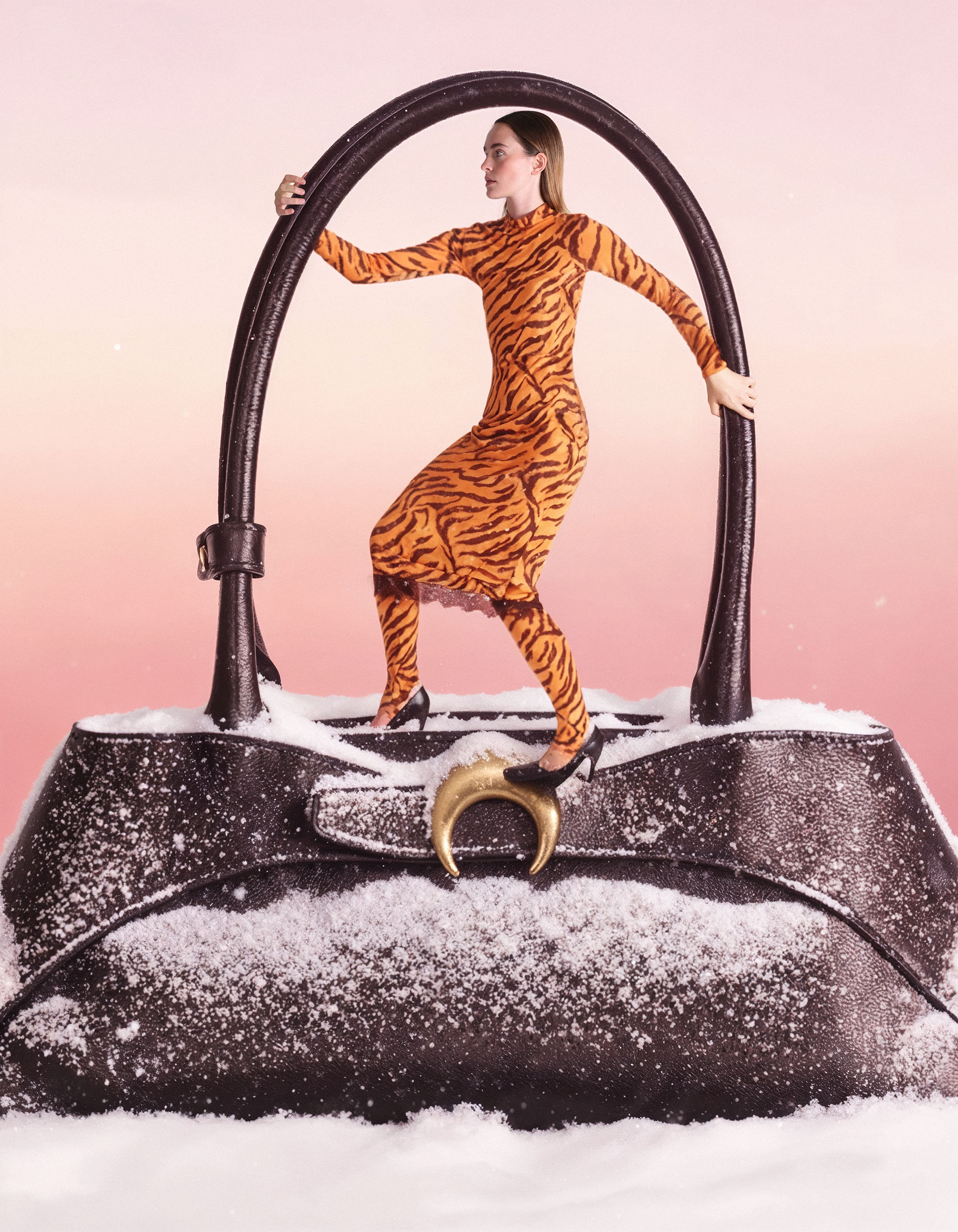 Fashion editorial shot of a woman in a tiger-print dress striking a pose on top of a snow-covered black leather purse with gold hardware.