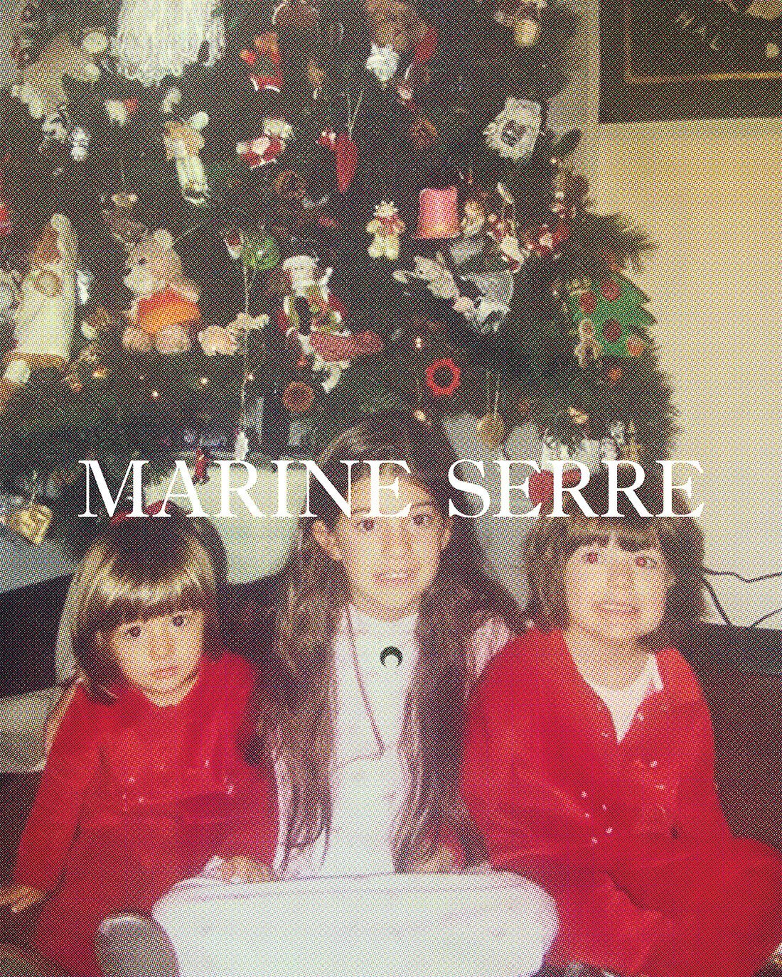 Three young girls pose in front of a decorated Christmas tree. Two are wearing red, and one is wearing white in an indoor setting.