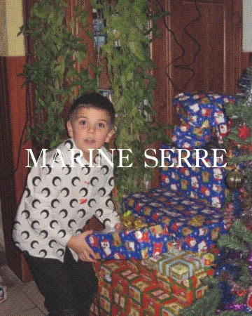 A child holds a Christmas present near a Christmas tree. The kid is wearing a white shirt with a black crescent moon pattern.