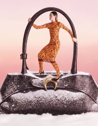 Model wearing orange tiger-print dress standing inside giant black handbag handle set in snowy landscape