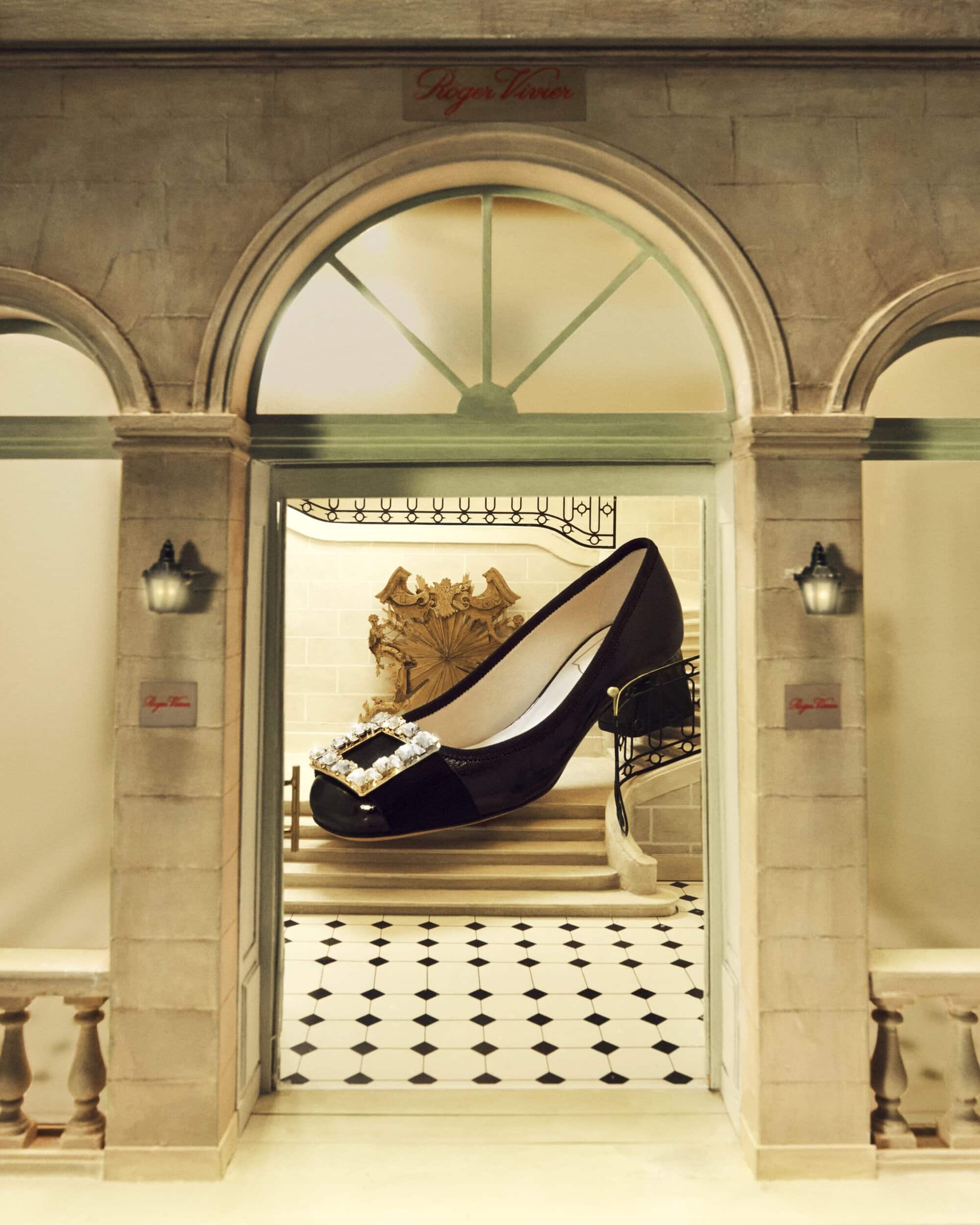 Oversized black Roger Vivier pump with a rhinestone buckle is showcased in front of an elegant marble staircase and black-and-white tiled floor.