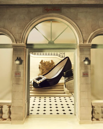 Oversized black Roger Vivier pump with a rhinestone buckle is showcased in front of an elegant marble staircase and black-and-white tiled floor.