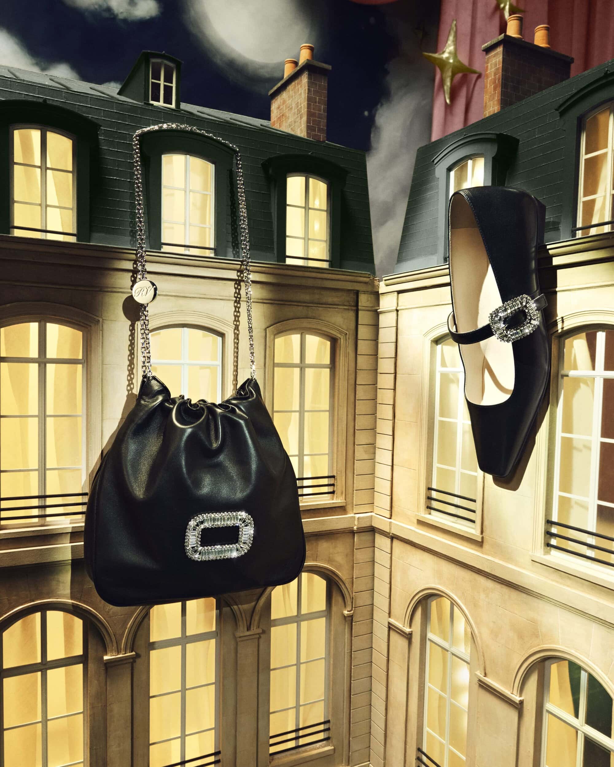 Elevated view of a black bucket bag with a chain strap and a black, jeweled shoe, suspended in front of illuminated building windows.